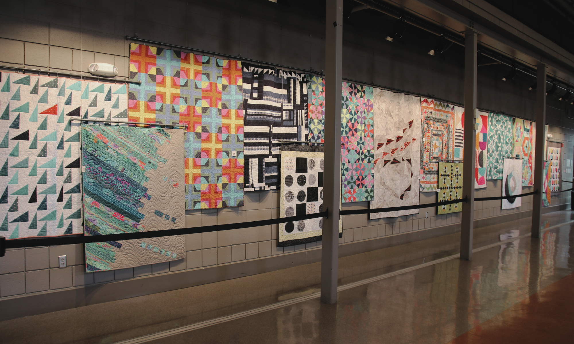 a variety of modern quilts hung for display on a painted concrete block wall in an art gallery