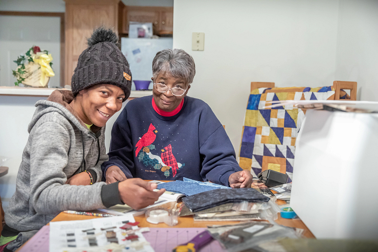 two Black women working together on a quilt project at a table, smiling at the camera